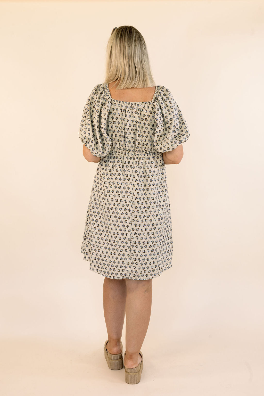 A woman viewed from the back wearing a knee-length dress with puff sleeves and an allover small floral print. The dress has a square neckline and a gathered waist.