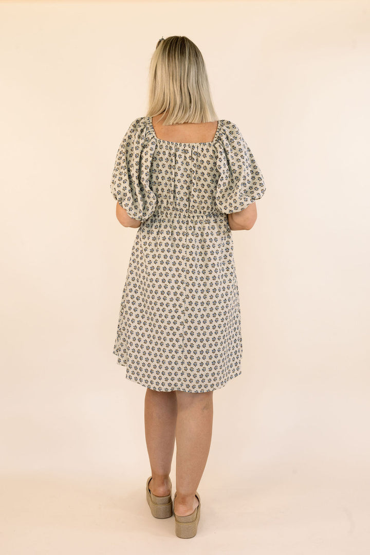 A woman viewed from the back wearing a knee-length dress with puff sleeves and an allover small floral print. The dress has a square neckline and a gathered waist.