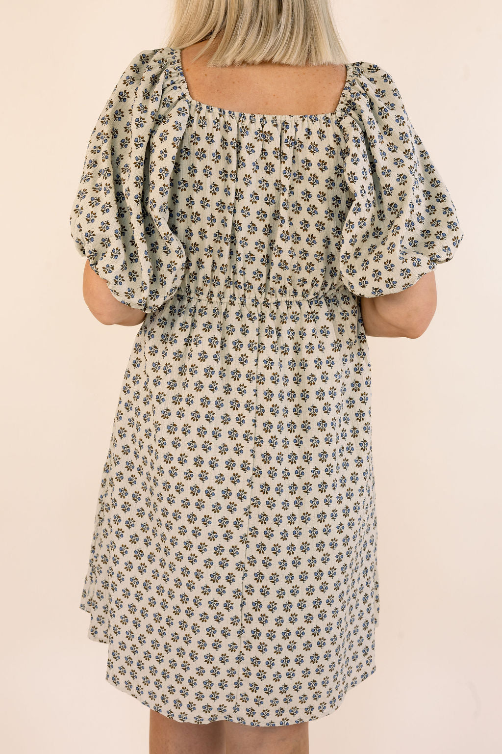 A woman wearing a short puff-sleeve dress with a small floral print, shown from the back against a plain light beige background.