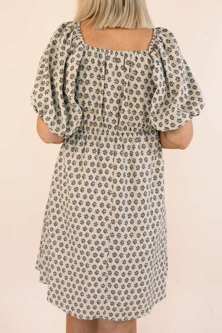 A woman wearing a short puff-sleeve dress with a small floral print, shown from the back against a plain light beige background.