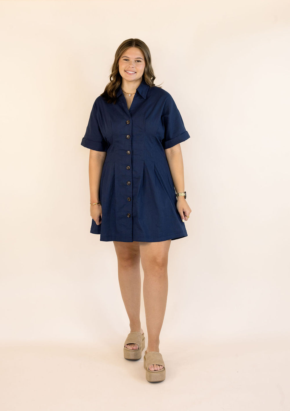 A woman modeling a short-sleeve navy blue button-up shirt dress with a shawl collar, standing against a plain beige studio background.