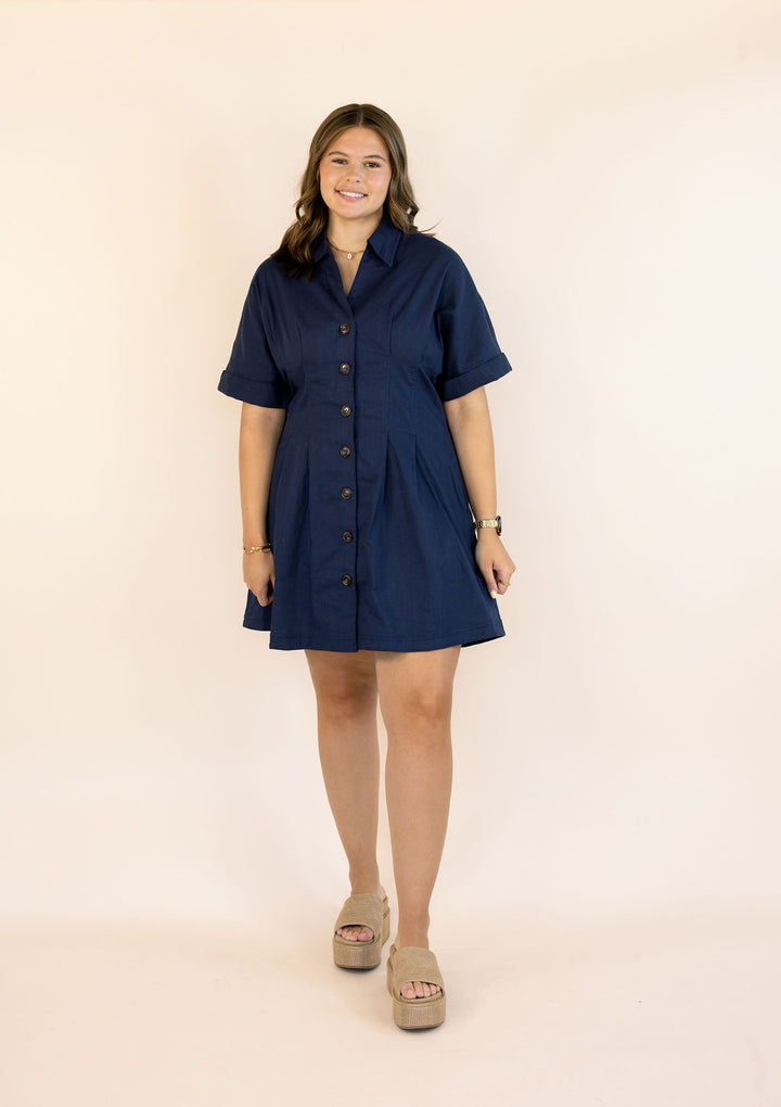 A woman modeling a short-sleeve navy blue button-up shirt dress with a shawl collar, standing against a plain beige studio background.