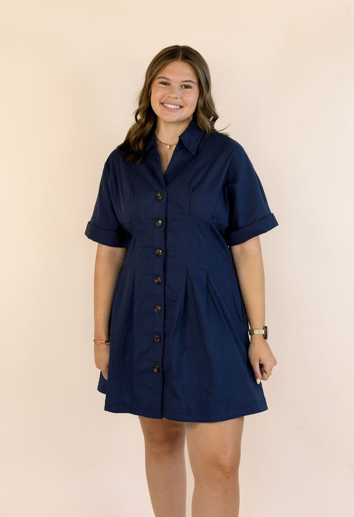 A woman modeling a navy blue short-sleeve button-front shirt dress against a solid light beige background.
