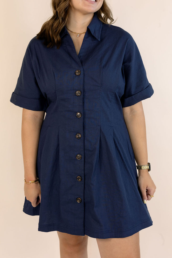 A woman wearing a navy blue short-sleeve button-front shirt dress with a pointed collar and pleated skirt, shown against a plain light beige background.