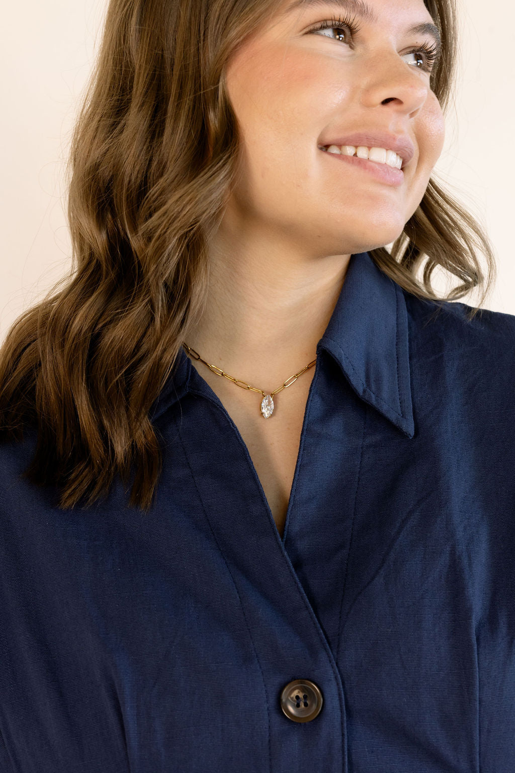 A close-up of a woman wearing a navy blue button-down shirt and a delicate gold necklace with a small clear pendant, set against a plain light beige background.