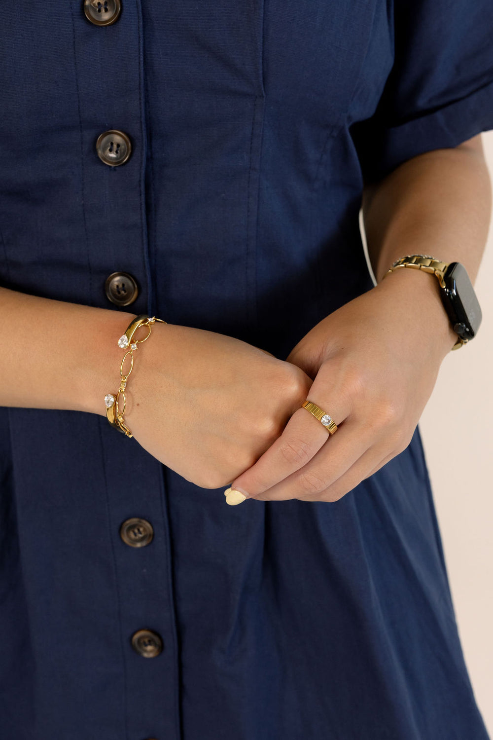 Close-up of a person wearing gold jewelry, including a delicate gold chain bracelet with small white stones, a gold ring, and a gold watch with a black band, paired with a navy blue button-up dress.