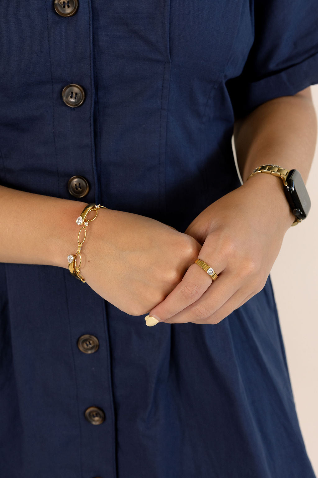 Close-up of a person wearing gold jewelry, including a delicate gold chain bracelet with small white stones, a gold ring, and a gold watch with a black band, paired with a navy blue button-up dress.