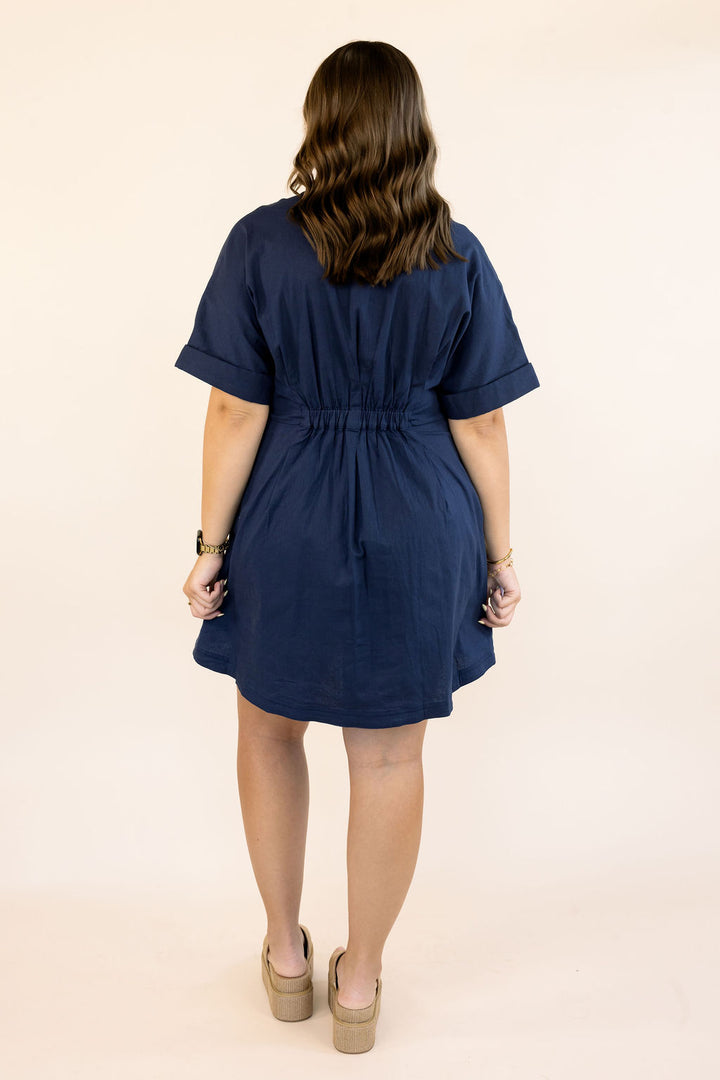 A woman viewed from behind wearing a navy blue short-sleeve dress with a gathered waist and beige platform heels, standing against a plain light beige studio background.