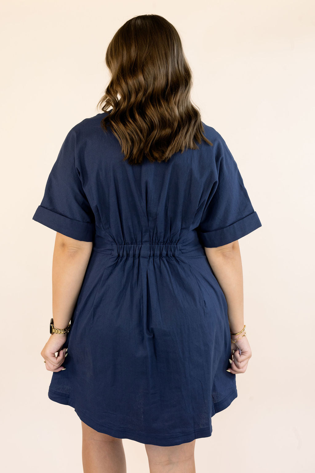 A woman viewed from behind wearing a navy blue short-sleeve dress with a gathered waist, set against a plain beige background.