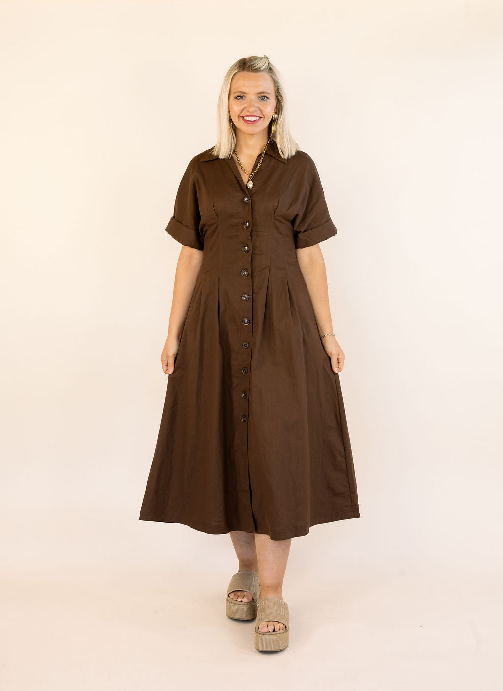 A woman modeling a brown short-sleeve midi dress with a button-down front, standing against a plain off-white studio background.
