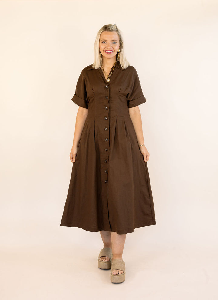 A woman modeling a brown short-sleeve midi dress with a button-down front, standing against a plain off-white studio background.
