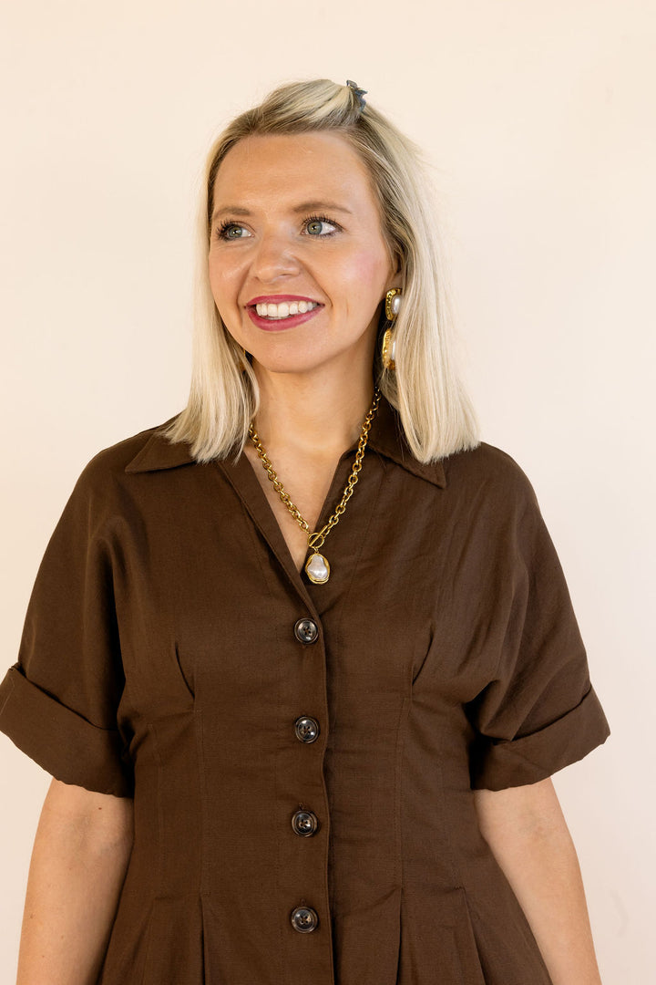 A smiling blonde woman wearing a brown short-sleeve button-front dress, standing against a plain light beige background.