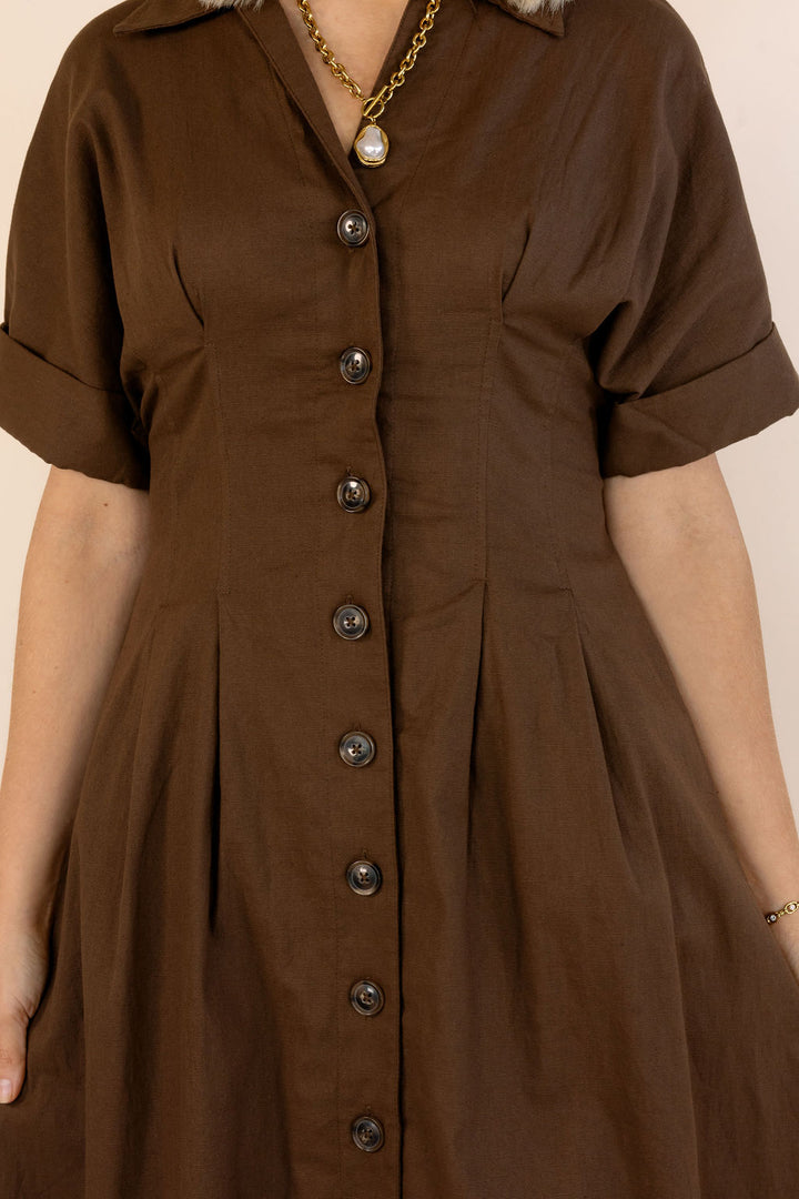 A close-up of a person wearing a brown short-sleeve button-front dress with a collar and pleated skirt, against a plain light beige background.