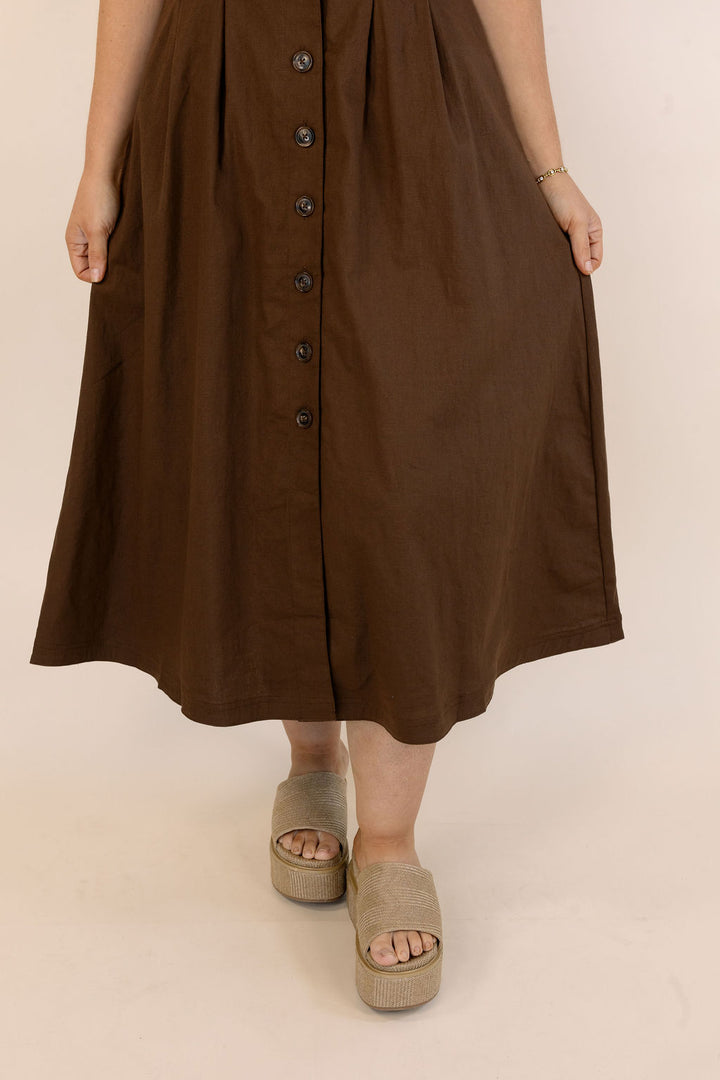 A person wearing a brown midi dress with a button-down front and pleated skirt, paired with beige platform sandals, shown against a plain beige studio background.