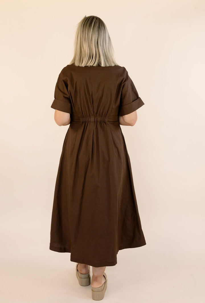 A woman viewed from behind wearing a brown short-sleeve midi dress with a gathered waist, standing against a plain beige studio background.