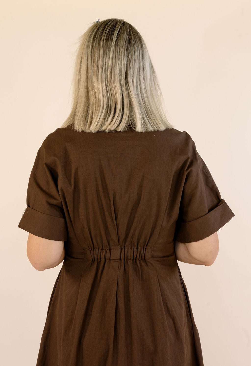A woman viewed from behind wearing a dark brown short-sleeve dress with rolled cuffs and a gathered waist, against a plain beige background.