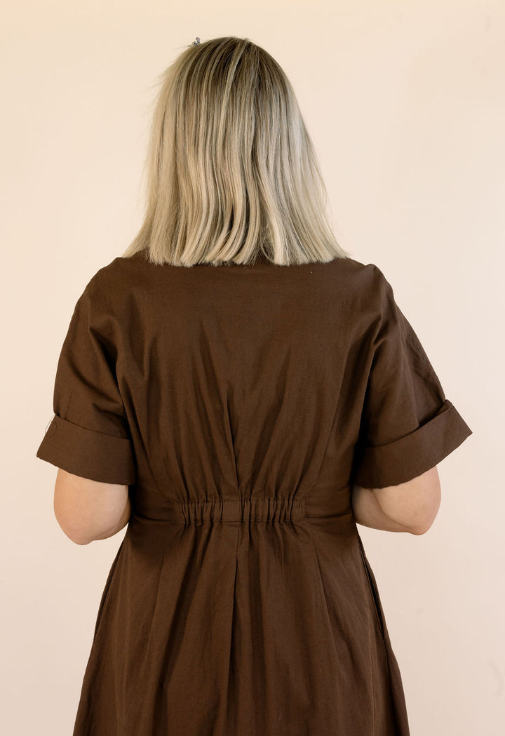 A woman viewed from behind wearing a dark brown short-sleeve dress with rolled cuffs and a gathered waist, against a plain beige background.