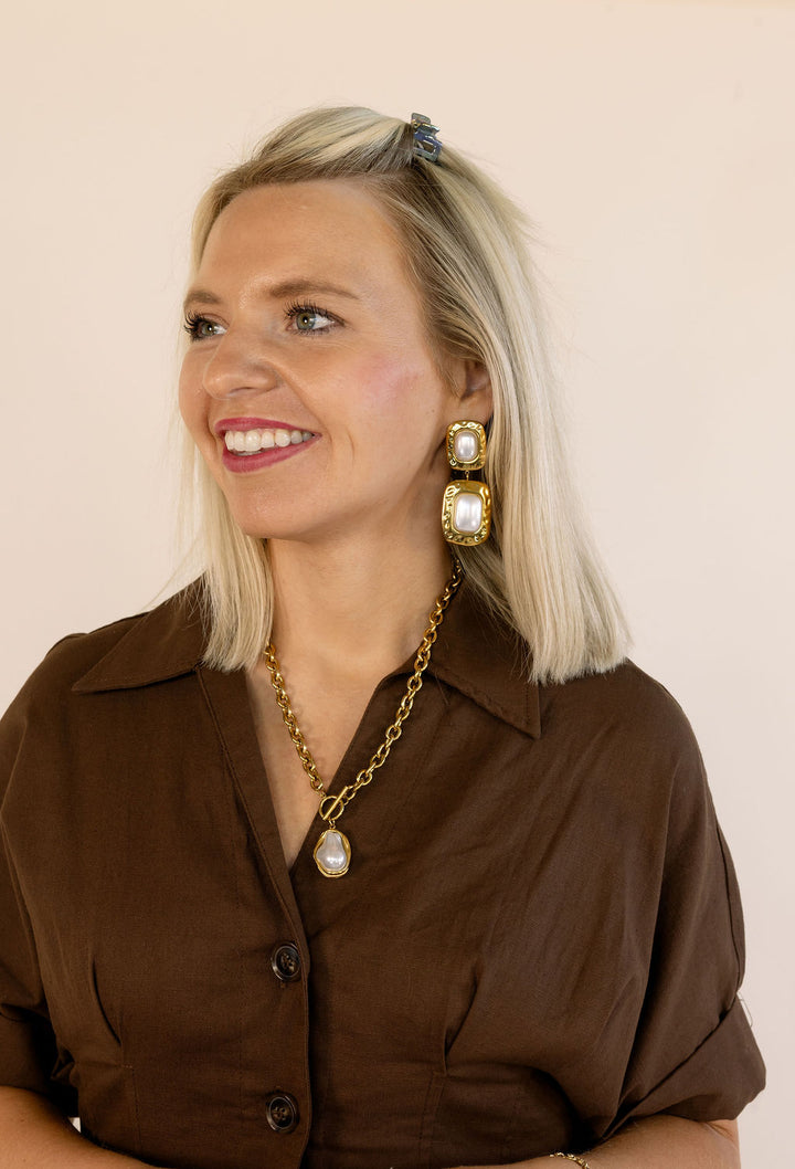 A smiling blonde woman wearing gold statement earrings with white pearl-like centers, a matching gold chain necklace with a teardrop pearl pendant, and a brown collared dress against a plain beige background.