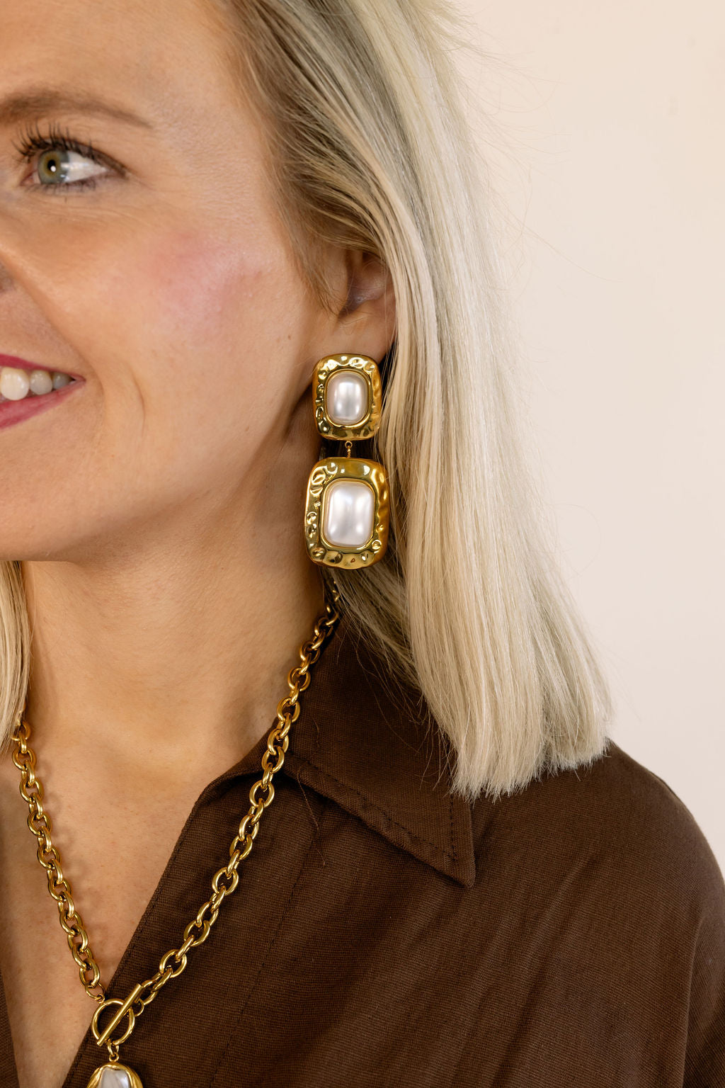 A close-up of a woman wearing a gold drop earring with two stacked rectangular pearl-like stones, along with a gold chain necklace and a brown top against a plain light beige background.