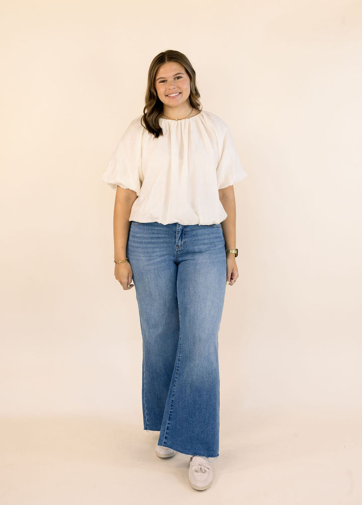 A woman modeling a cream-colored short-sleeve bubble blouse and wide-leg blue jeans against a solid beige studio backdrop.
