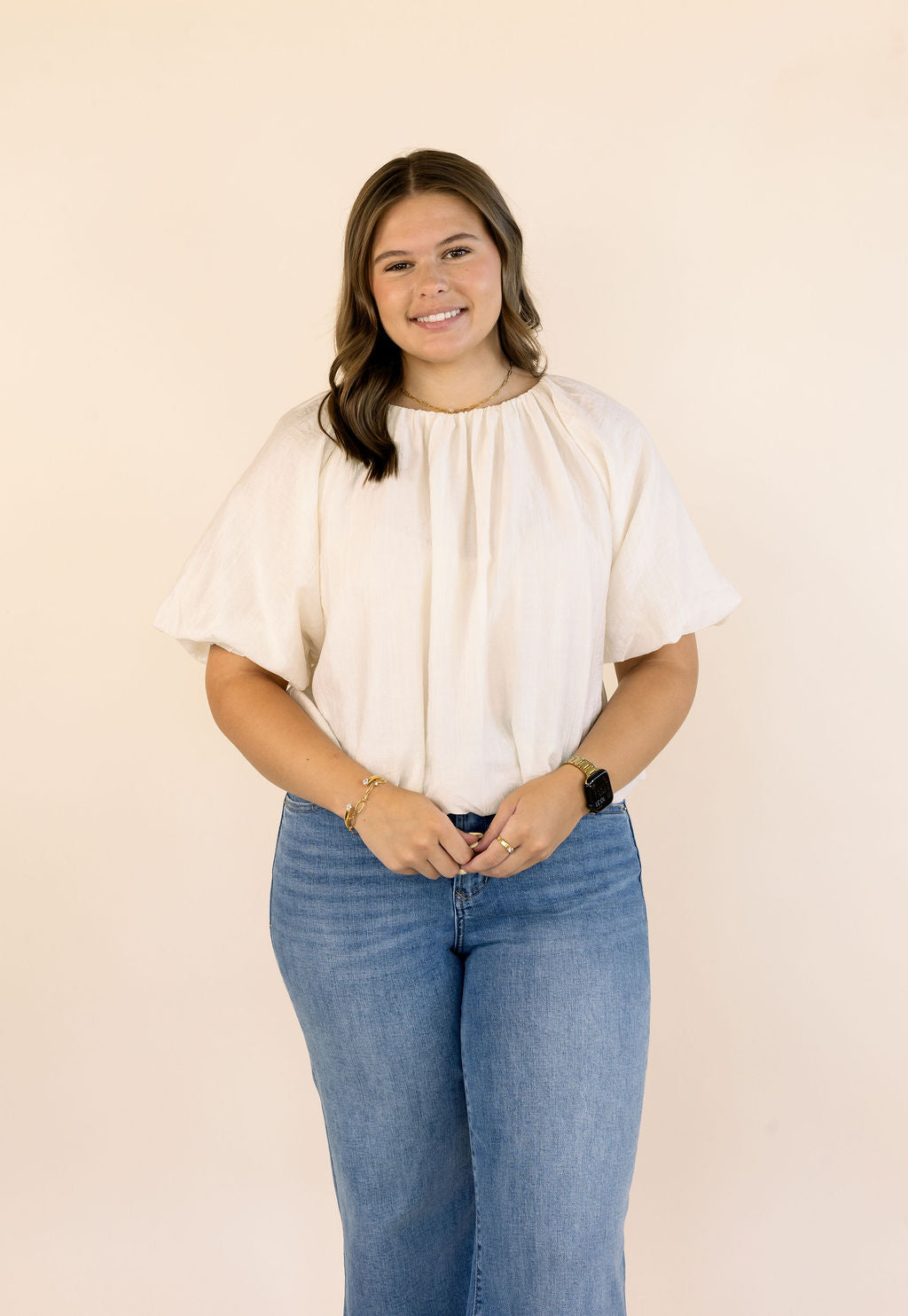 A woman modeling a cream-colored short-sleeve blouse with a gathered, pleated neckline, paired with blue jeans, standing against a solid light beige background.