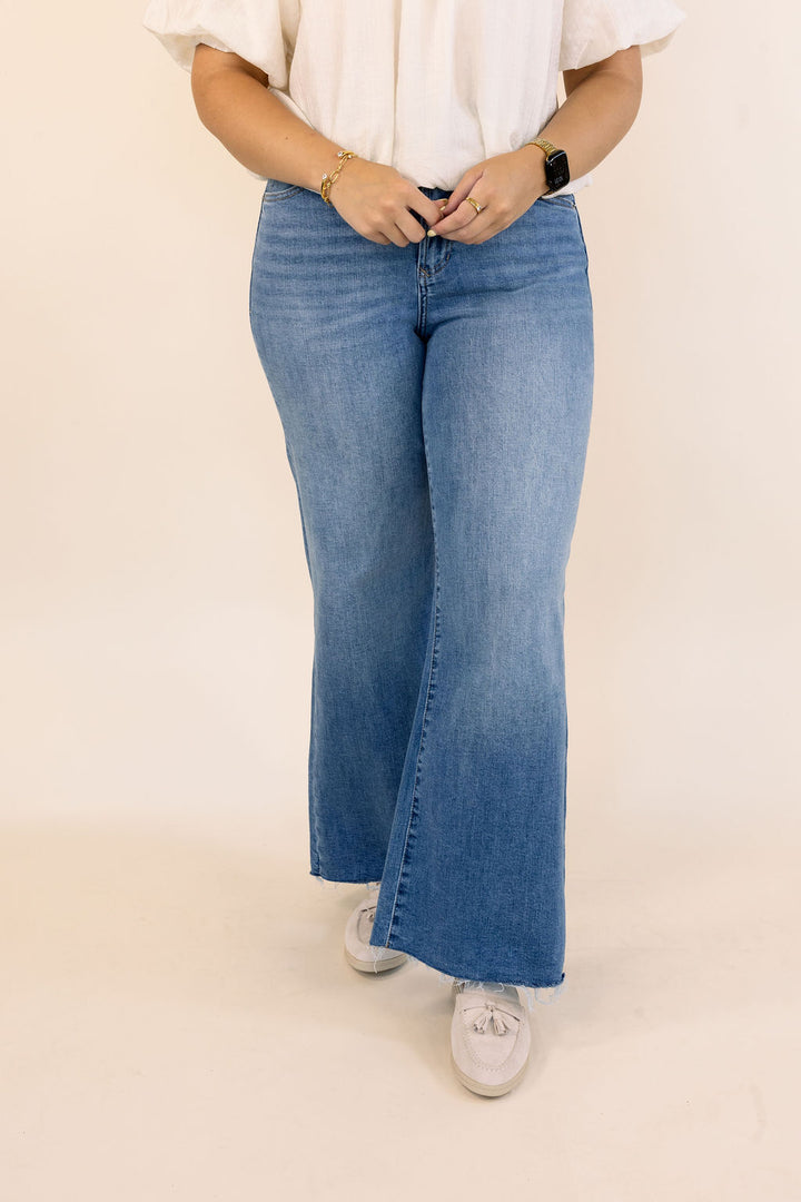 A person wearing medium-wash blue flare jeans with a white blouse and white mules, standing against a plain beige studio background.