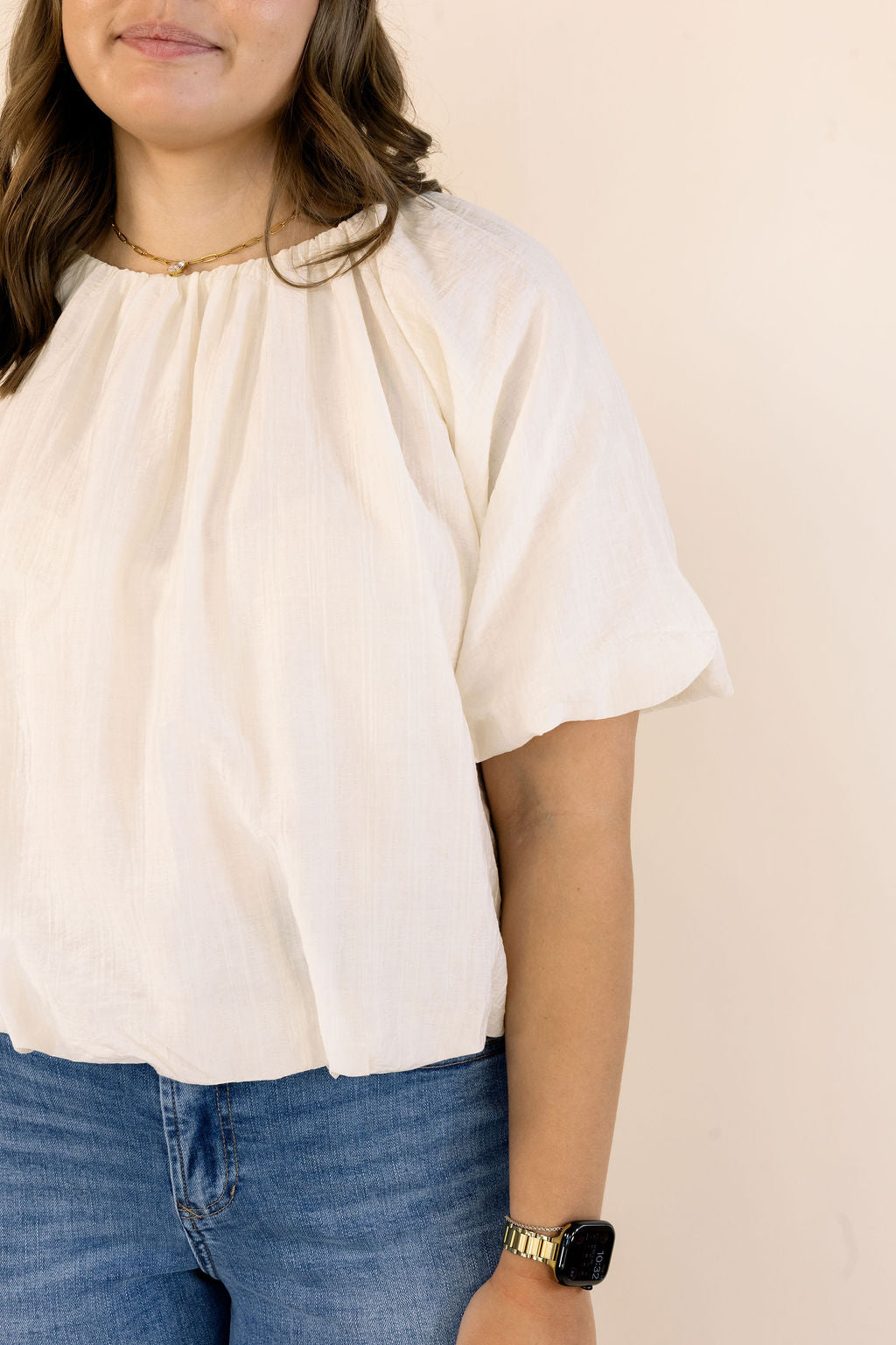 A woman wearing a cream-colored short-sleeve blouse with a gathered, pleated neckline, paired with blue jeans, shown against a plain beige background.