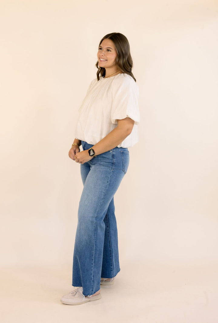 A woman modeling a cream short-sleeve bubble blouse with puff sleeves and blue wide-leg jeans against a plain beige studio background.