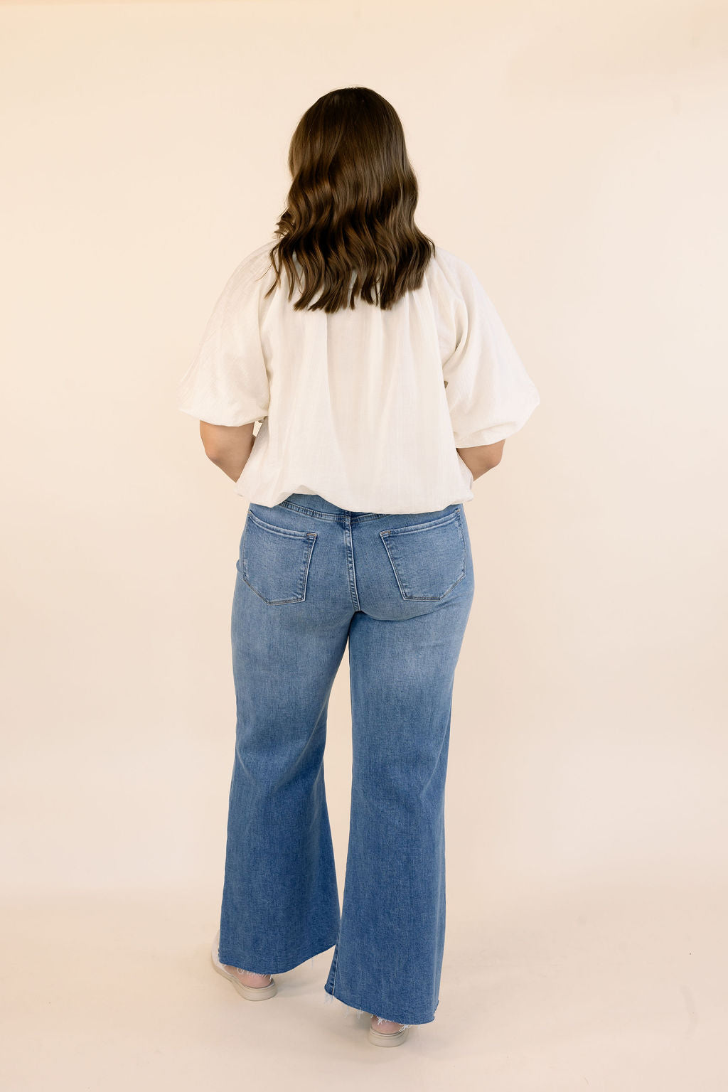 A woman viewed from behind wearing a cream short-sleeve bubble hem top and blue wide-leg jeans against a plain beige studio background.