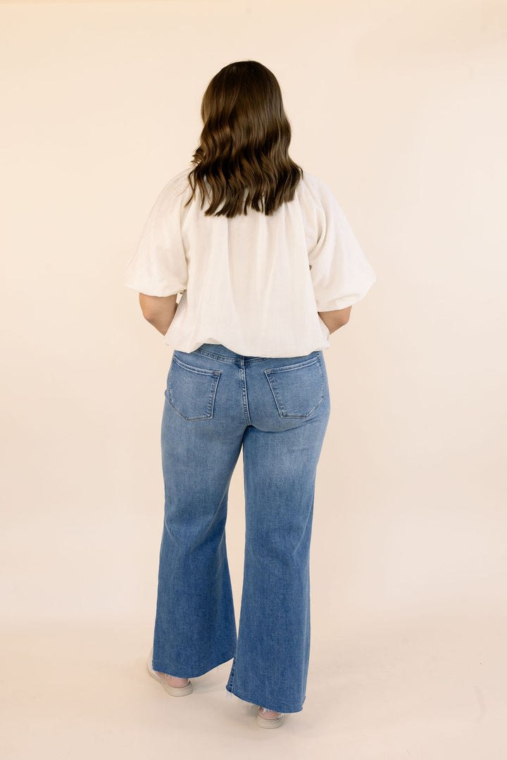 A woman viewed from behind wearing a cream short-sleeve bubble hem top and blue wide-leg jeans against a plain beige studio background.