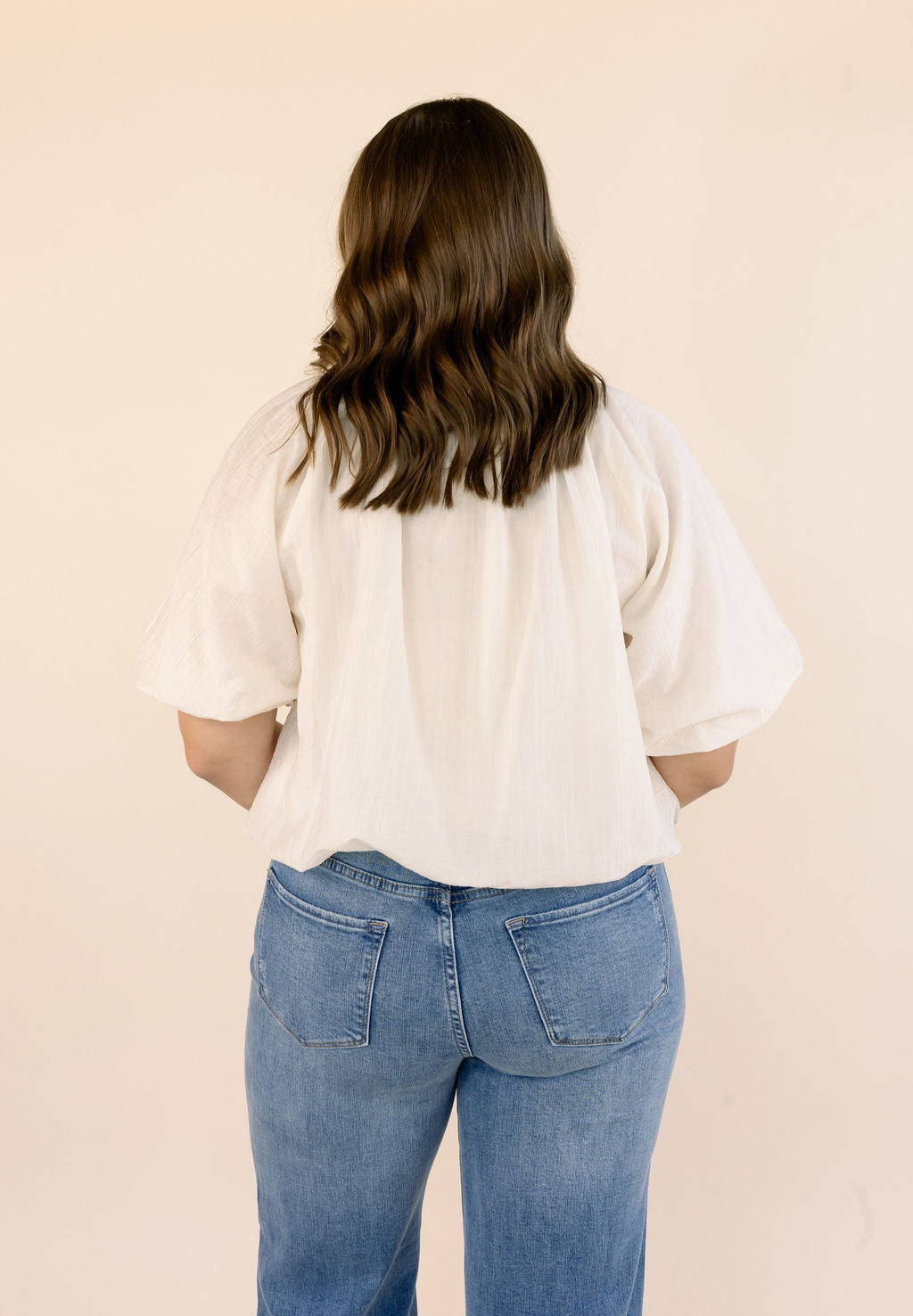 A woman viewed from behind wearing a cream-colored short-sleeve blouse and blue jeans, standing against a solid light beige background.