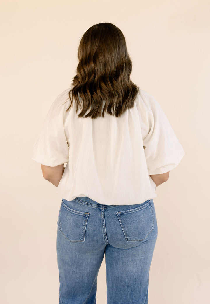 A woman viewed from behind wearing a cream-colored short-sleeve blouse and blue jeans, standing against a solid light beige background.