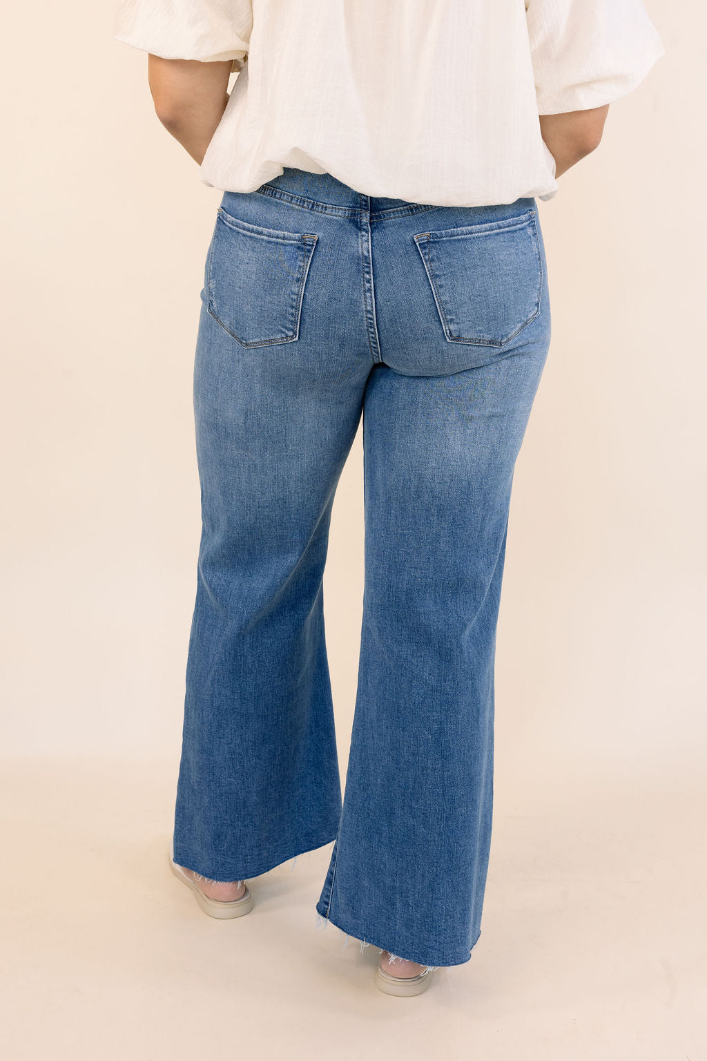 A person shown from the back wearing blue wide-leg denim jeans and a white blouse against a plain light beige studio background.
