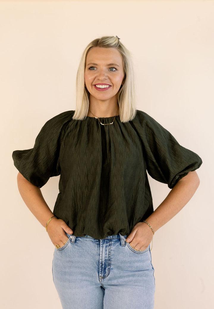 A smiling blonde woman wearing a dark olive green bubble blouse with voluminous puff sleeves and light-wash blue jeans, standing against a plain beige background.