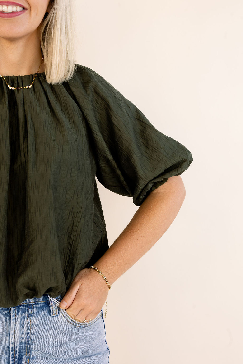 A woman wearing a dark olive green crinkled short-sleeve blouse with light-wash blue jeans, shown from the chest down against a plain light beige background.