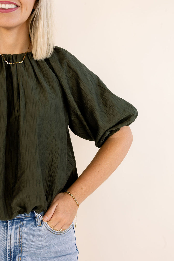 A woman wearing a dark olive green crinkled short-sleeve blouse with light-wash blue jeans, shown from the chest down against a plain light beige background.