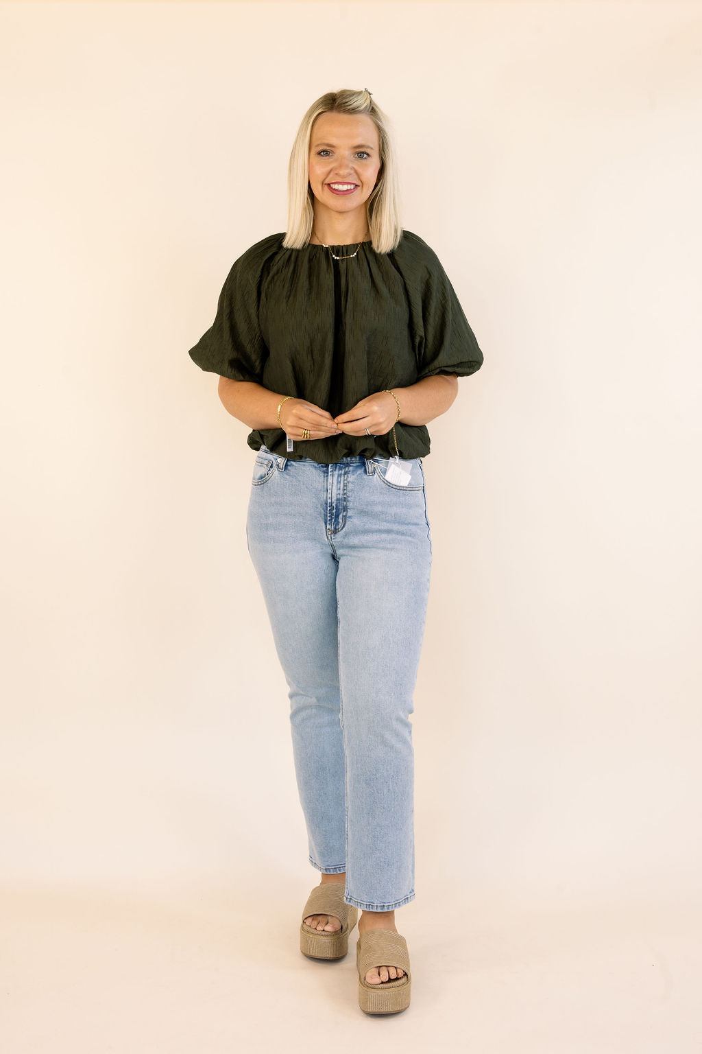 A woman modeling a dark olive green short-sleeve bubble blouse with puff sleeves, paired with light-wash blue jeans and beige platform sandals, standing against a solid cream background.