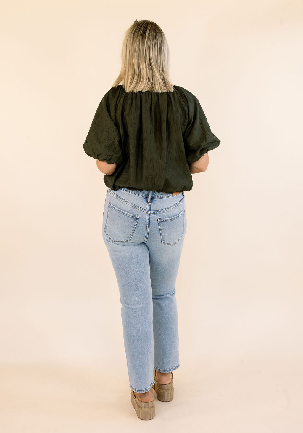 A woman viewed from behind wearing a dark olive green puff-sleeve blouse and light-wash blue jeans, standing against a plain beige studio background.