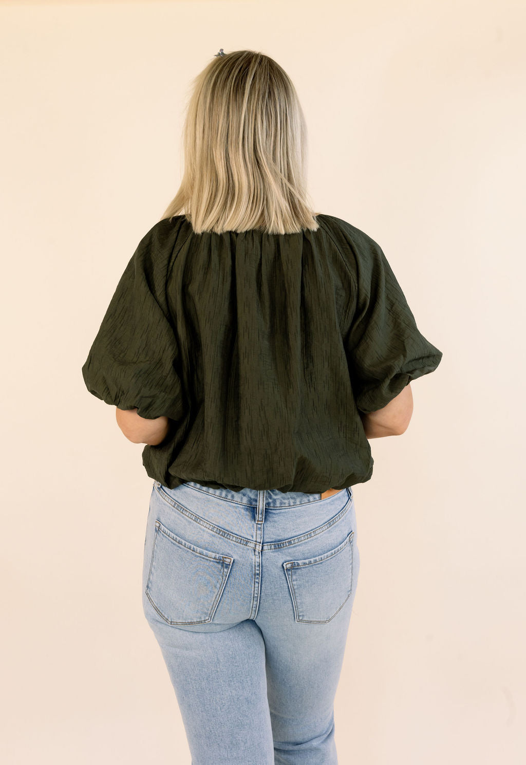 A woman viewed from behind wearing an olive green short-sleeve blouse with voluminous puff sleeves and light blue jeans, standing against a plain beige background.