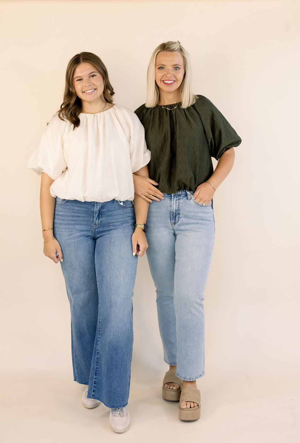 Two women standing side-by-side modeling different styles of light-wash blue jeans and matching tops against a solid cream background. The woman on the left wears a cream bubble hem top and the woman on the right wears the same shirt in olive.