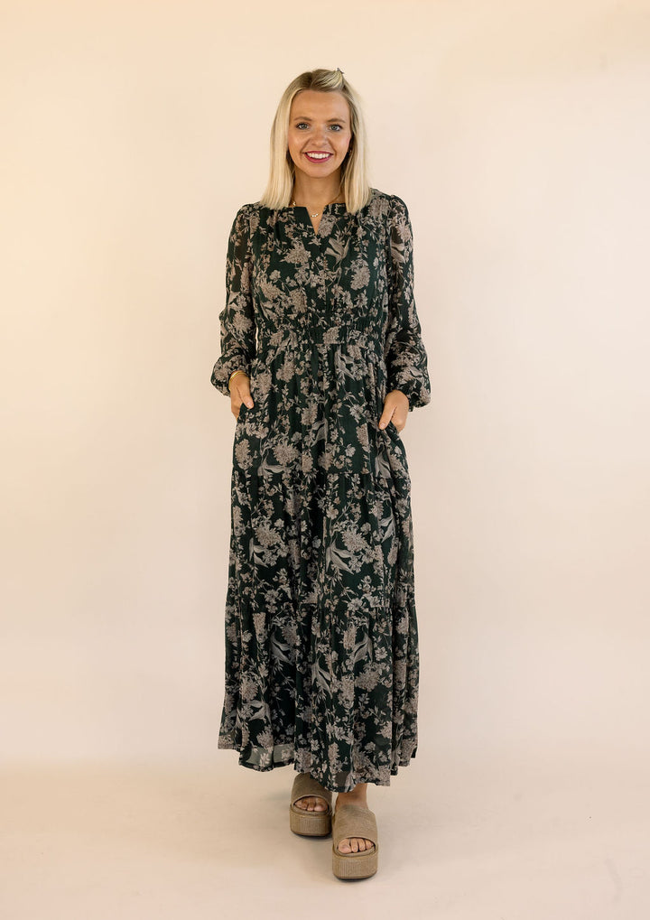 A woman modeling a long-sleeved maxi dress with a dark green and beige floral pattern, standing against a plain beige studio background.