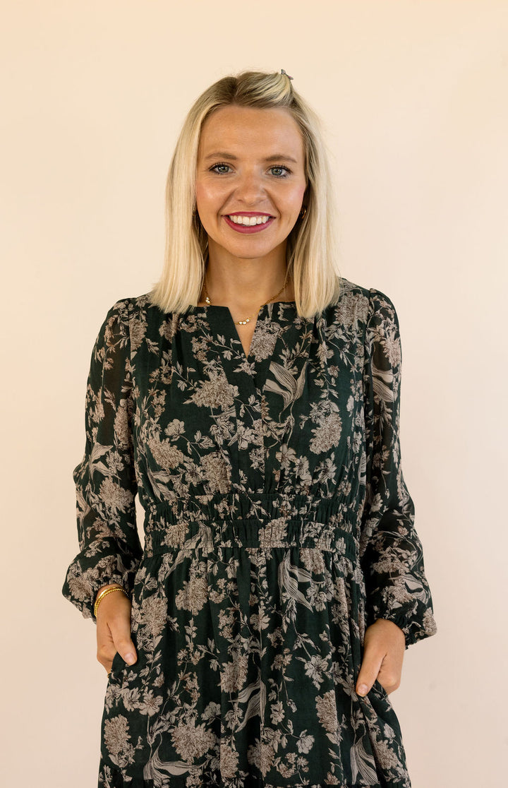 A smiling blonde woman wearing a dark green long-sleeve dress with a beige floral pattern, standing against a solid light beige background.