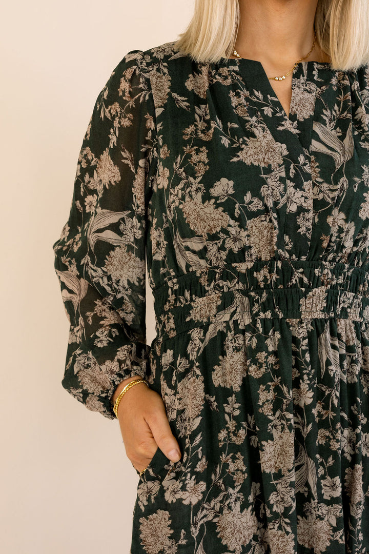 A close-up of a woman wearing a dark green long-sleeve dress with a beige floral pattern, featuring a smocked waist and a split neckline.