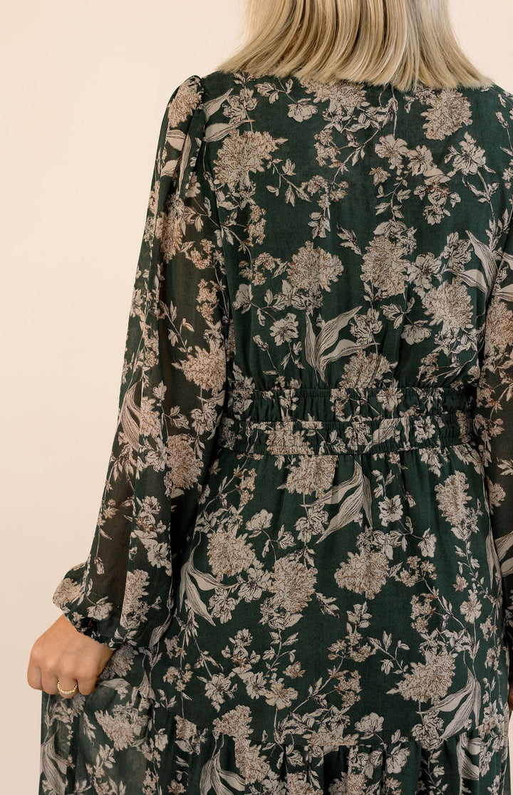 Back view of a dark green floral print midi dress featuring long sleeves and a gathered elastic waist detail.