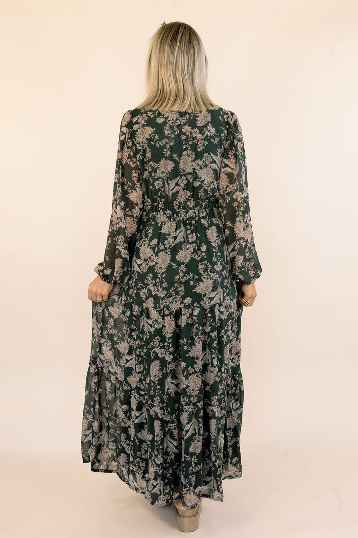 A woman viewed from behind wearing a long-sleeved, floor-length maxi dress with a dark green and beige floral pattern, set against a plain beige studio background.