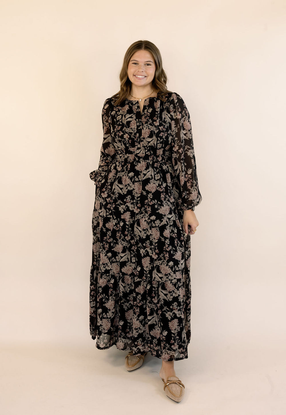 A woman modeling a long-sleeved black maxi dress with a beige floral pattern, standing against a plain beige studio background.