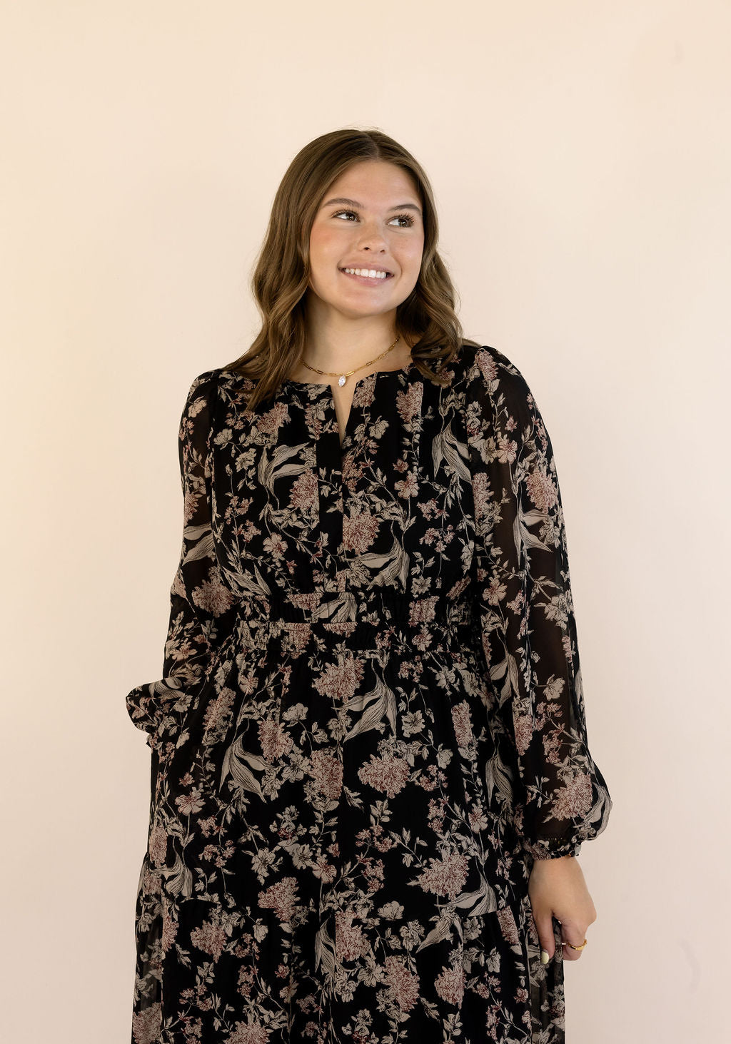 A woman modeling a long-sleeve black dress with a beige floral pattern, standing against a solid pale beige background.