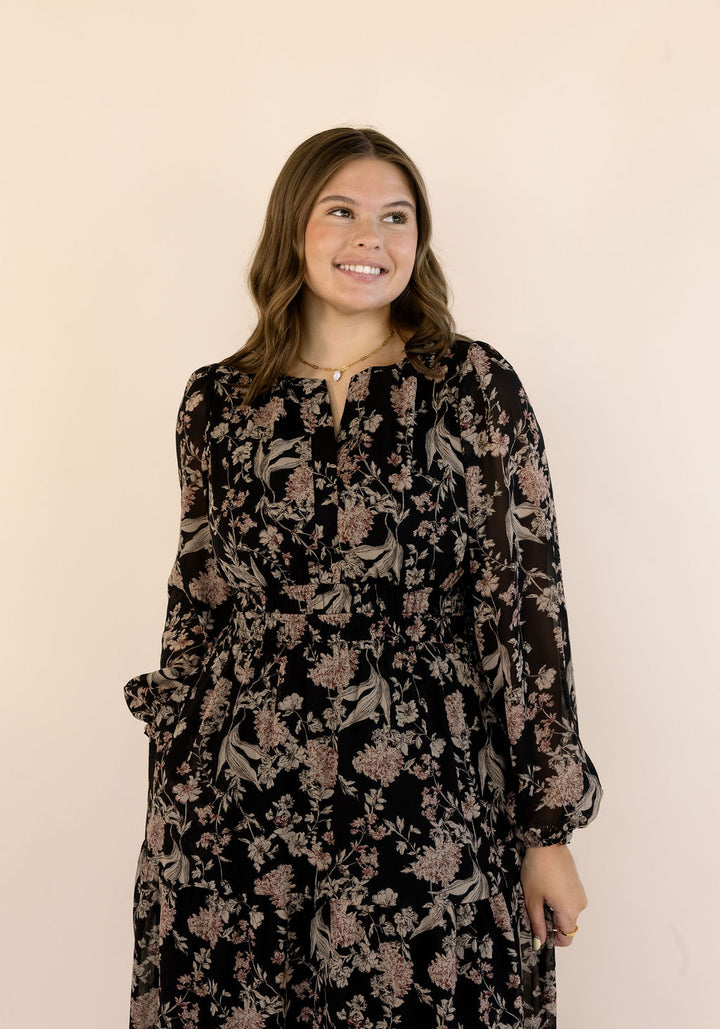 A woman modeling a long-sleeve black dress with a beige floral pattern, standing against a solid pale beige background.