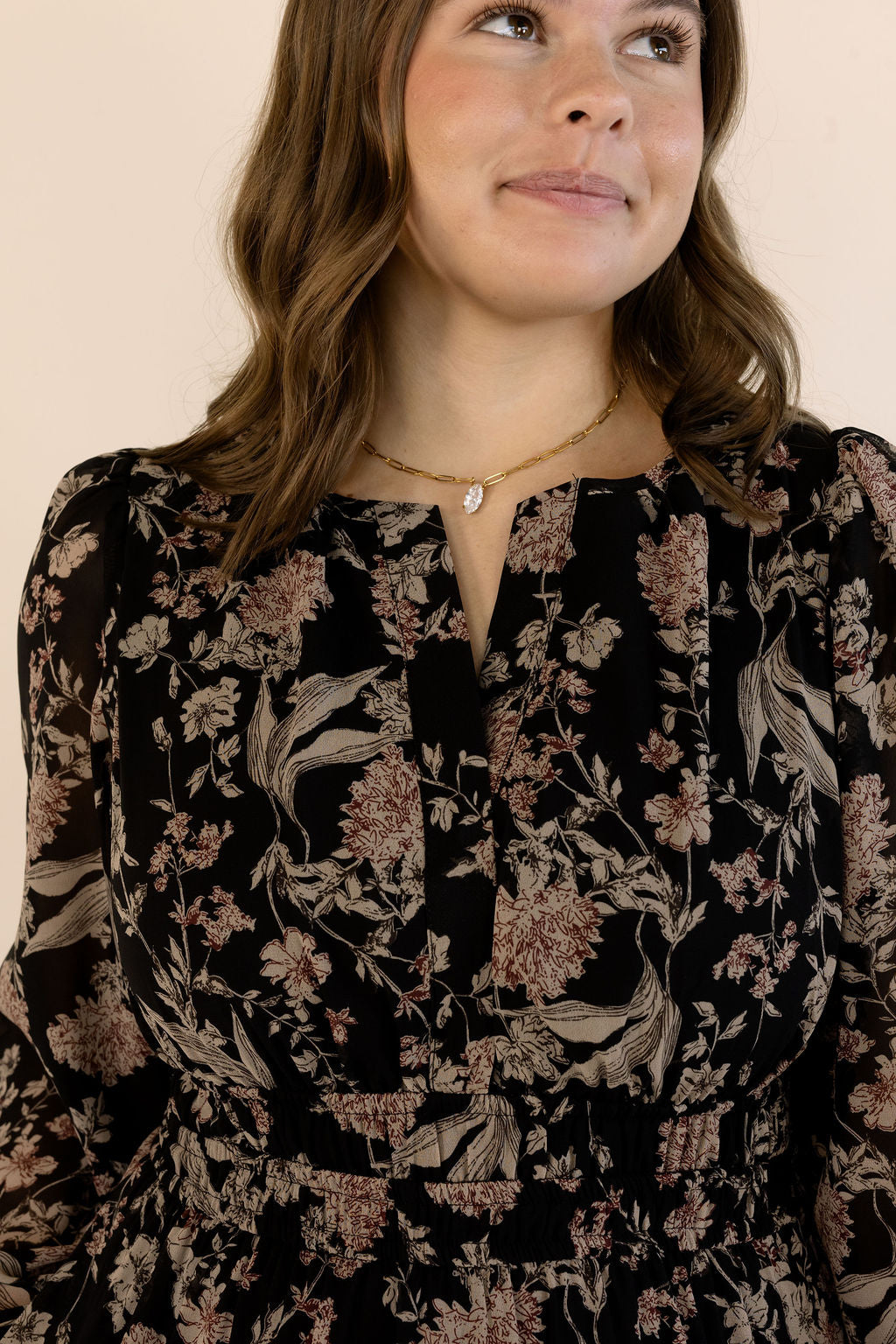 A woman wearing a black long-sleeve dress with a dusty rose and beige floral pattern, paired with a delicate gold necklace, set against a plain beige background.