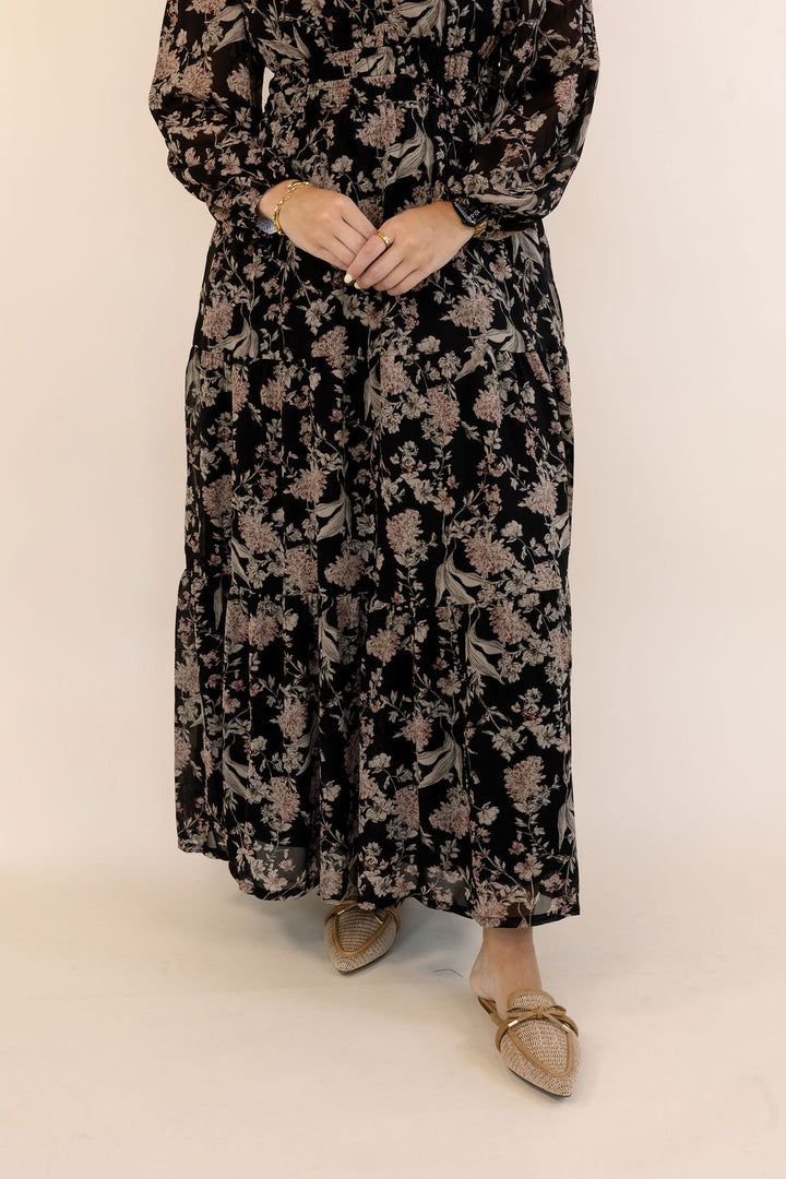 A woman wearing a long-sleeved black maxi dress with a dusty rose and beige floral pattern, paired with beige flat shoes, standing against a plain off-white background.